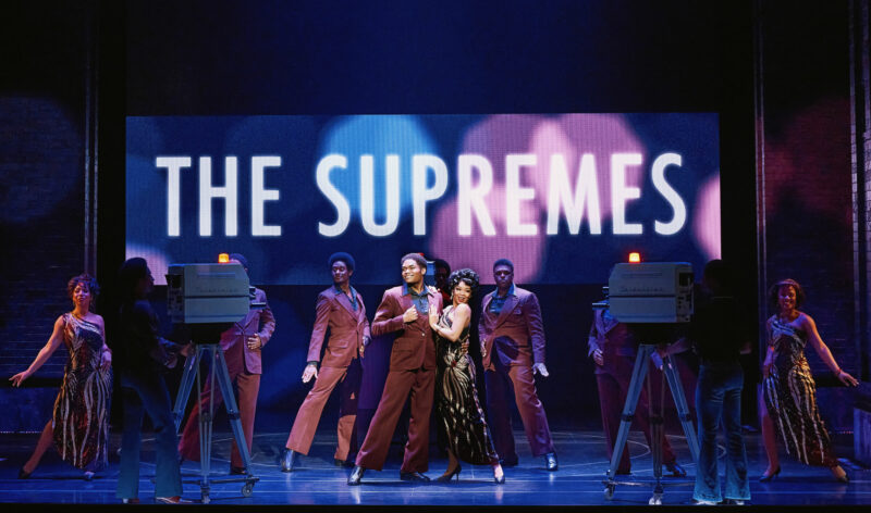 The National Touring Company of Ain’t Too Proud. A group of people performing in front of cameras and a big sign that reads “The Supremes”.